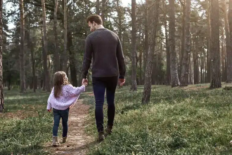 A father and a young child walking hand in hand through a forest, seen from behind. Therapy for third culture kids and multicultural families.