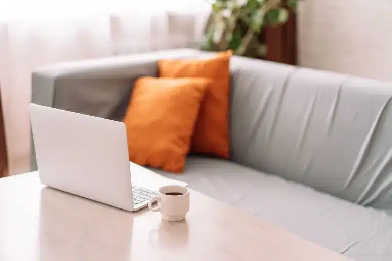 A laptop and a cup on a table in front of a sofa with orange cushions. Counseling for children, teens, adults and families near Tigard, Oregon.
